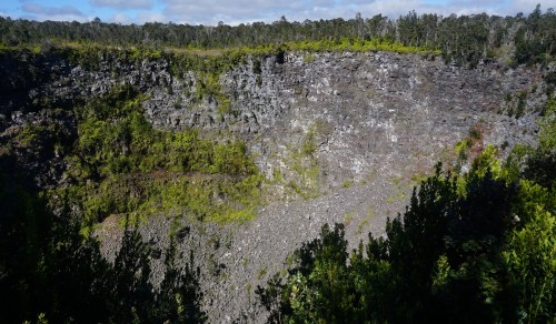 A rocky volcanic crater surrounded by lush greenery and trees under a partly cloudy sky.