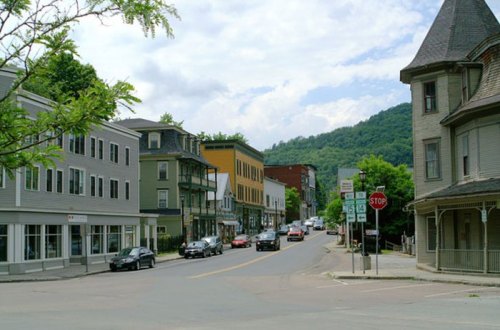 A quaint street scene with historic buildings, parked cars, and a backdrop of green hills under a cloudy sky.