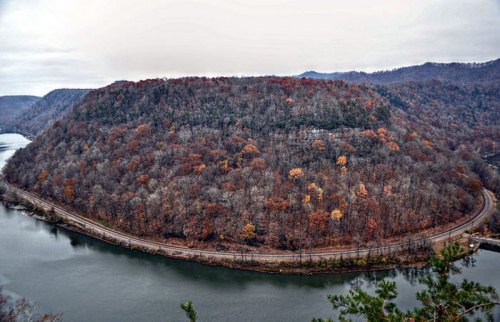 A scenic view of a river curving around a wooded hill, showcasing autumn foliage under a cloudy sky.