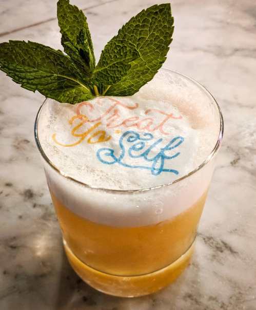 A cocktail topped with foam and mint leaves, featuring the text "Treat Yo Self" in colorful lettering.