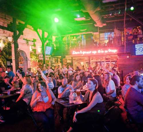 A lively crowd enjoying an event at a bar, with colorful lights and a neon sign in the background.