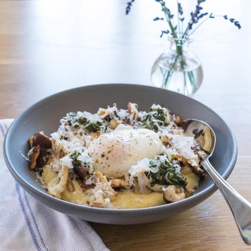 A bowl of creamy polenta topped with sautéed greens, mushrooms, a poached egg, and grated cheese, with a vase of flowers in the background.