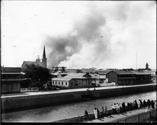 A historic black-and-white photo showing a river, buildings, and smoke rising in the background, with people along the bank.