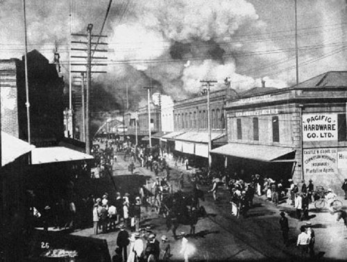 Historic street scene with crowds, buildings, and smoke in the background, depicting a bustling urban area.