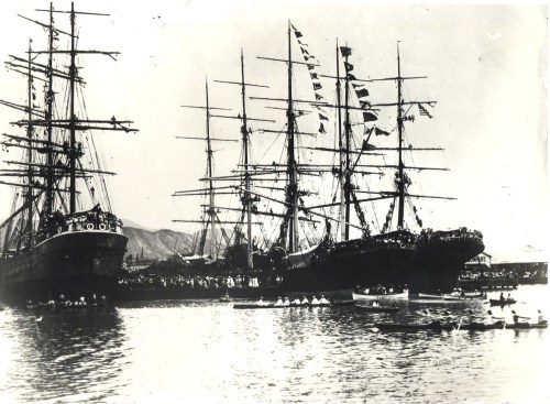 Historic black-and-white photo of tall ships docked, with small boats nearby and a crowd on the shore.