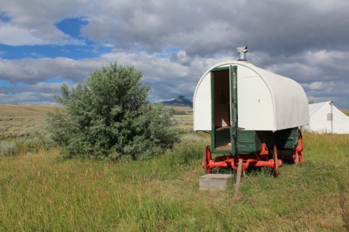 A vintage white and green wagon on red wheels, set in a grassy field with a cloudy sky and distant mountains.
