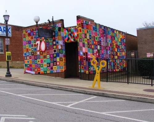 Colorful patchwork mural covers a building's exterior, with a large pair of scissors sculpture in front.