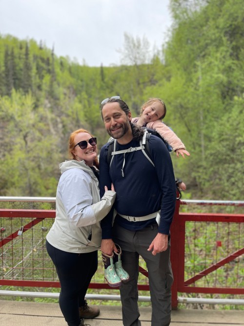 A smiling family poses on a bridge in nature, with a woman, a man, and a child on the man's back. Green trees in the background.