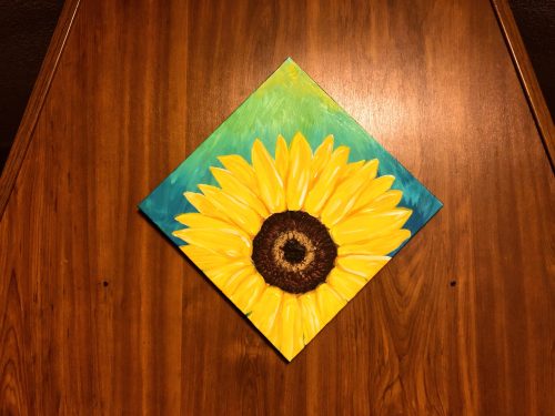 A vibrant painting of a sunflower on a wooden background, featuring bright yellow petals and a dark center.