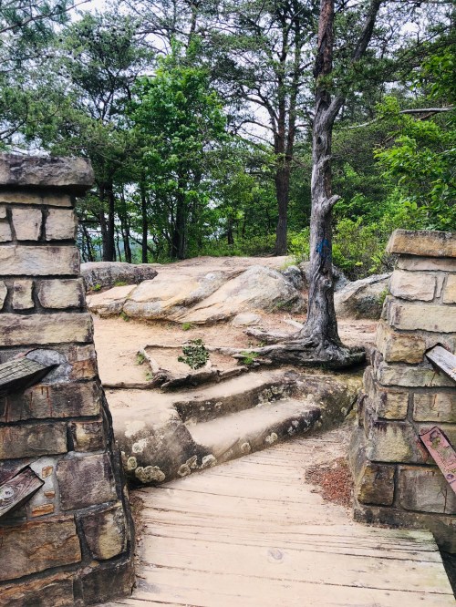 A stone archway leads to a rocky path surrounded by trees and greenery in a natural setting.