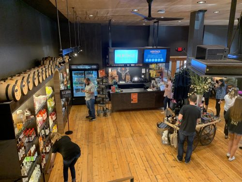 A spacious store with wooden floors, featuring shelves of products, a bar area, and customers browsing and shopping.