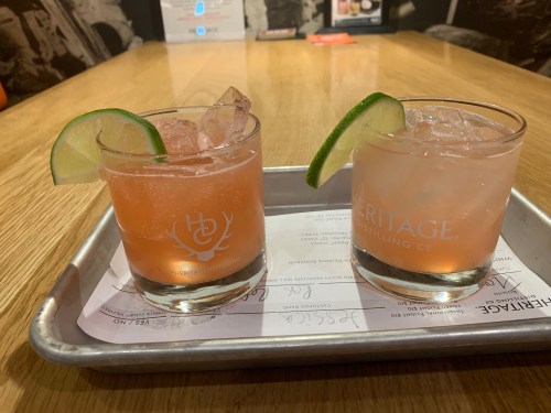 Two cocktails on a tray, one pink with a lime wedge and one clear with ice, set on a wooden table.
