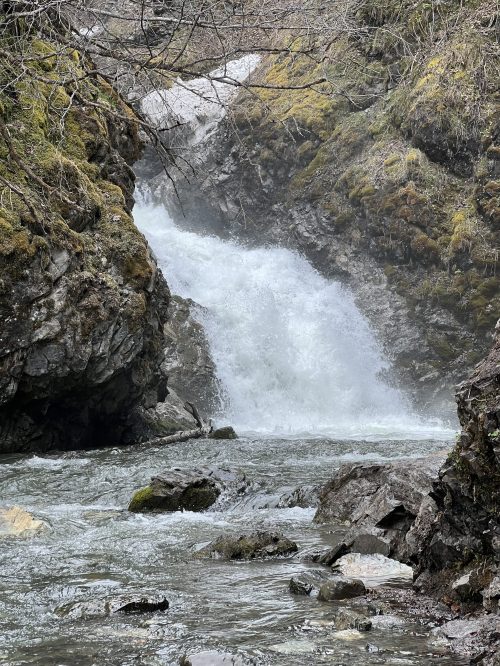 A cascading waterfall flows between rocky cliffs, surrounded by moss and greenery in a serene natural setting.