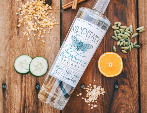 A bottle of organic gin surrounded by herbs, spices, and fresh fruits on a wooden surface.
