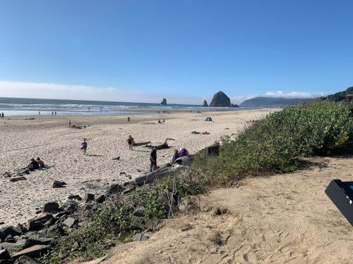 A sandy beach with people relaxing, rocky formations in the distance, and clear blue skies.