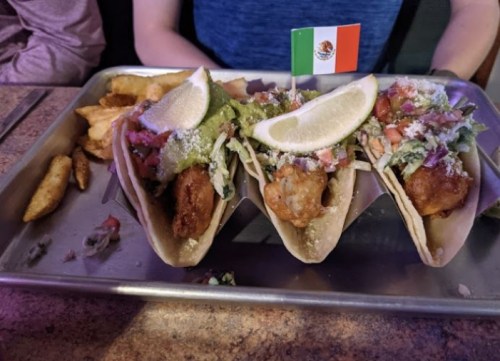 Three fish tacos topped with guacamole, pico de gallo, and a lime wedge, served on a metal tray with fries.