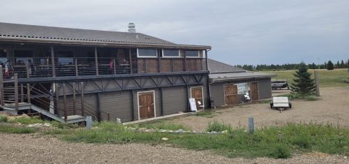 A rustic building with a balcony, wooden doors, and a gravel area, surrounded by greenery and a cloudy sky.