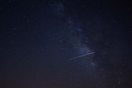 A starry night sky filled with bright stars and a visible streak of a meteor or satellite across the Milky Way.