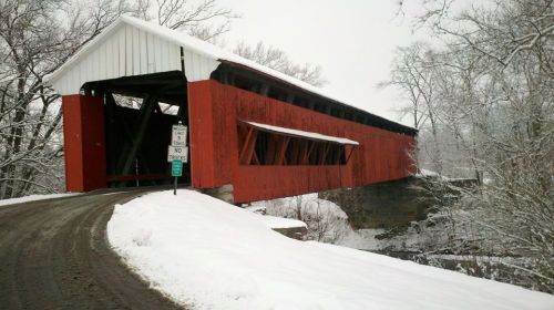 A red covered bridge in winter, surrounded by snow-covered trees and a gravel road leading to it.