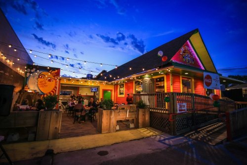 A colorful restaurant exterior at dusk, adorned with string lights and a mural, with diners visible inside.