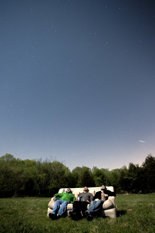 Three people relax on a couch in a field, gazing up at a starry night sky.