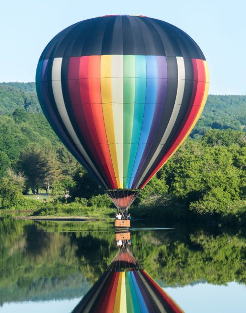 A colorful hot air balloon reflects on a calm lake, surrounded by lush greenery and trees under a clear blue sky.