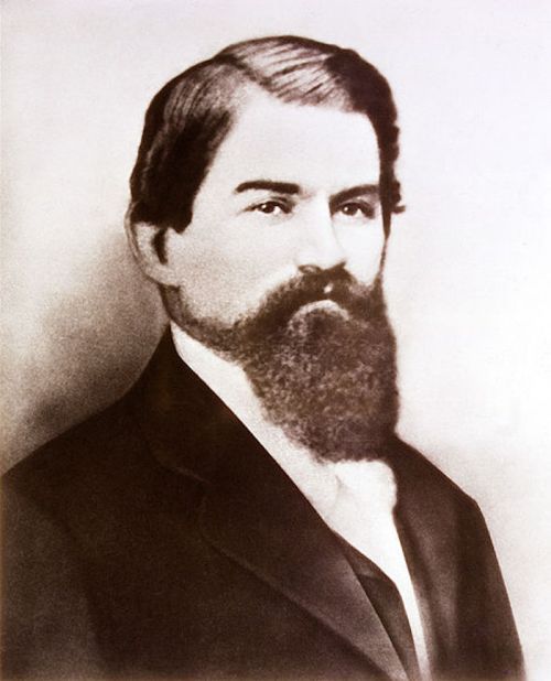 A black-and-white portrait of a man with a beard, wearing a suit and tie, looking directly at the camera.