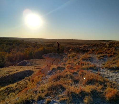 A person stands on a rocky ledge overlooking a vast landscape at sunset, with golden light illuminating the scene.