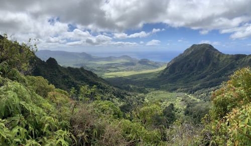 A panoramic view of lush green mountains and valleys under a partly cloudy sky, overlooking a coastal landscape.