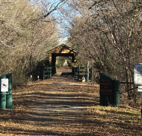 A wooden bridge leads through a tree-lined path, with signs indicating private property and no trespassing.