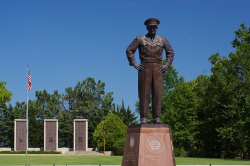 A bronze statue of a military officer stands in a park, with trees and memorial pillars in the background under a clear blue sky.