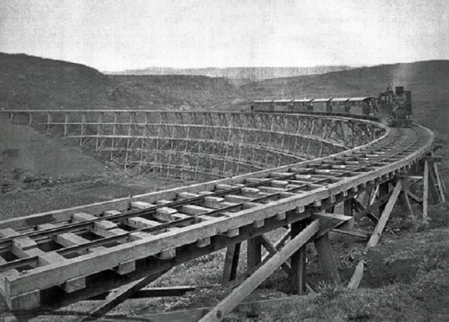 A vintage train on a curved wooden trestle bridge in a mountainous landscape.