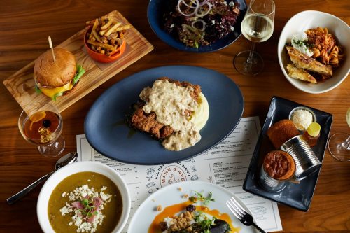 A spread of various dishes including fried chicken with gravy, a burger, pasta, and salads on a wooden table.