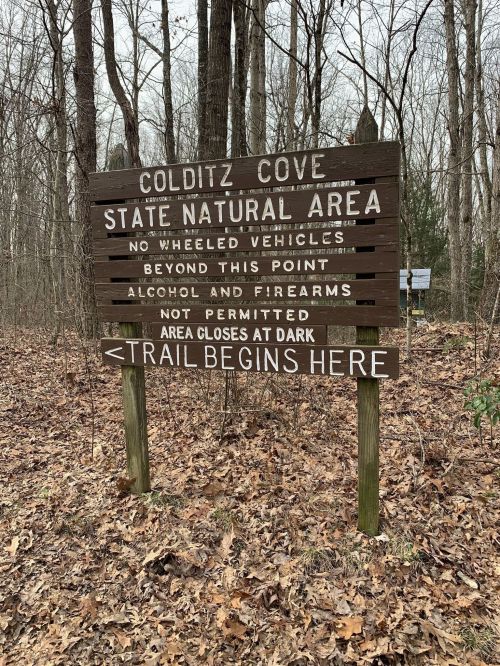 Sign at Colditz Cove State Natural Area indicating no vehicles, alcohol, or firearms allowed; trail begins here.