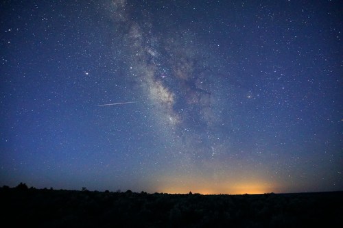 A stunning night sky filled with stars and the Milky Way, with a faint glow on the horizon and a shooting star.