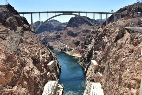 A view of a river flowing through rocky canyons, with a large arch bridge spanning above. Power stations are visible below.