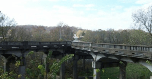 A view of a weathered concrete bridge surrounded by trees and greenery under a blue sky with scattered clouds.