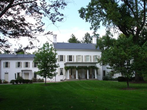 A large white house with green trees and a lawn, featuring a porch and multiple windows under a blue sky.