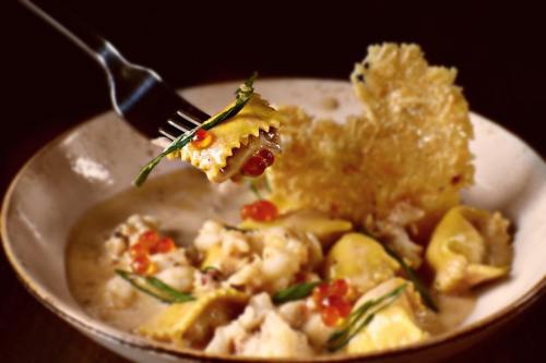 A fork holding a piece of ravioli above a bowl of creamy pasta with herbs and garnishes.
