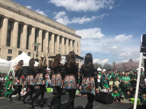A group of dancers in colorful costumes perform on stage at a lively outdoor event with a large crowd in green attire.