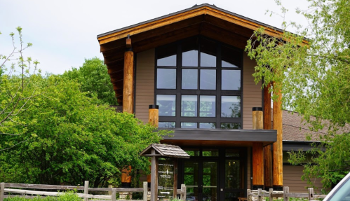 A modern building with large windows and wooden accents, surrounded by greenery and a wooden fence.