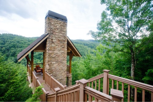 A wooden deck with a stone chimney overlooks a lush green valley and mountains under a cloudy sky.