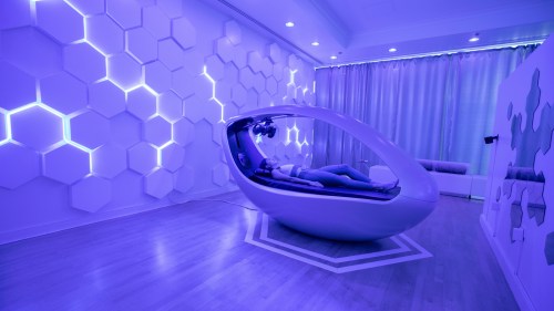 A person relaxes in a futuristic pod in a softly lit, purple-toned room with hexagonal wall designs.