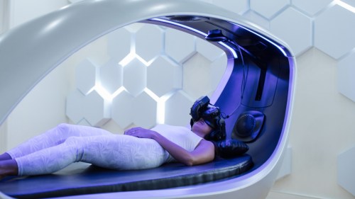 A person lies inside a futuristic VR pod, wearing a headset, in a modern, softly lit room with hexagonal patterns.