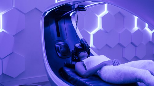 A person lies on a futuristic chair wearing a VR headset in a dimly lit, purple-toned room.