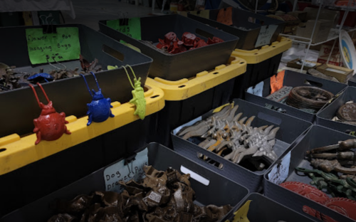 Colorful plastic bugs hang from a black and yellow bin, surrounded by various metal items in other bins.