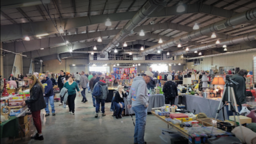 A bustling indoor market with many people browsing various vendor tables and colorful displays.
