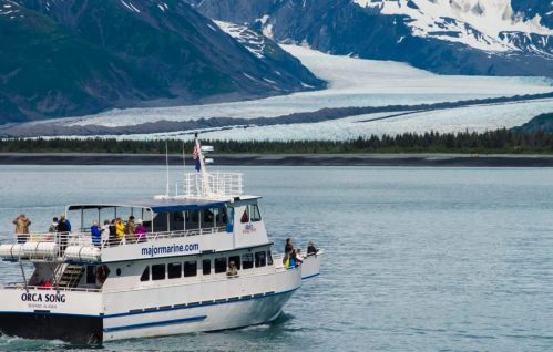 A tour boat named "Orca Song" sails near a glacier, surrounded by mountains and lush greenery.