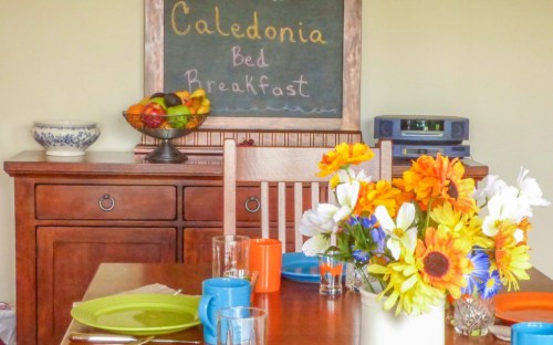 A cozy dining area with a wooden table set for breakfast, colorful flowers, and a chalkboard menu in the background.