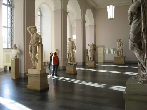 A spacious museum gallery featuring classical sculptures and visitors admiring the artwork. Natural light streams in.